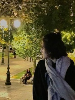 A person with dark hair and a scarf looks to the side in a park at night, with trees and a streetlamp in the background, their expression reflecting stories of strength and hope and love. | Support refugees across Europe in Greece, France, the UK and the Balkans