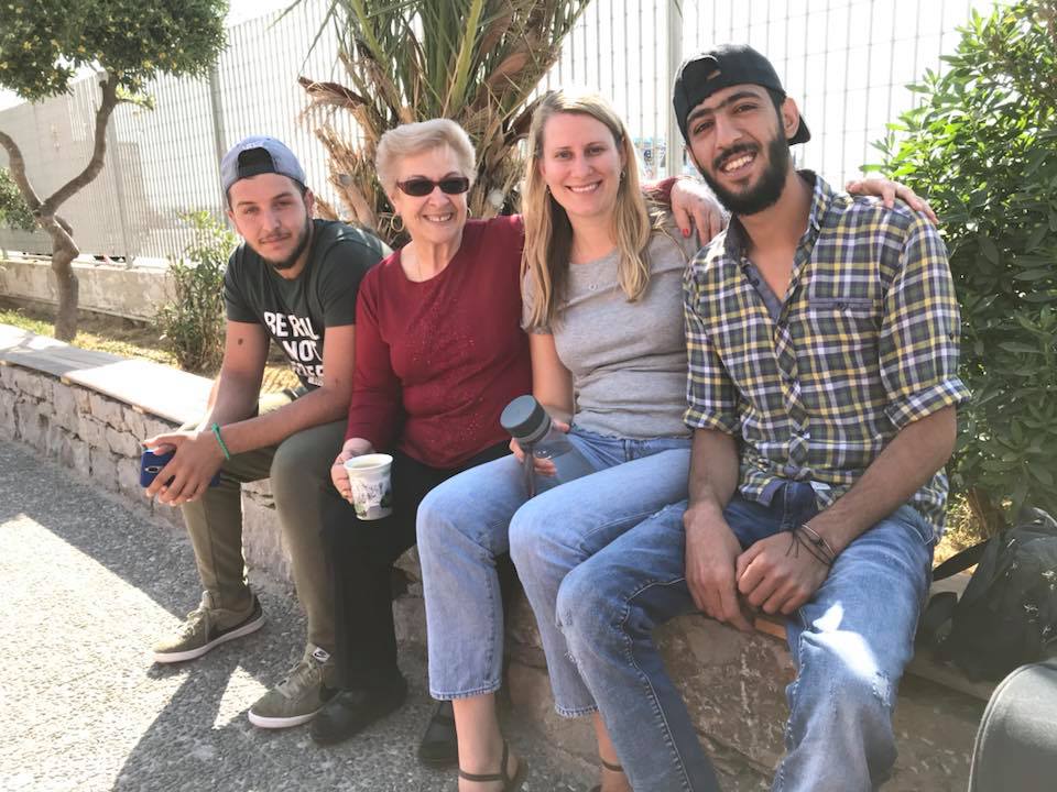 Four people sit on a low wall outdoors in Chios Island, Greece—smiling at the camera on Day 2. Two women in the middle hold cups, with two men seated on either side. | Support refugees across Europe in Greece, France, the UK and the Balkans