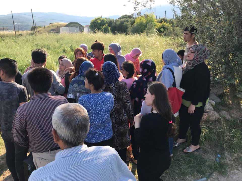 A group of people, many wearing headscarves, stand together outdoors in a grassy area on Chios Island, Greece, enjoying the sunny Day 2 of their journey. | Support refugees across Europe in Greece, France, the UK and the Balkans