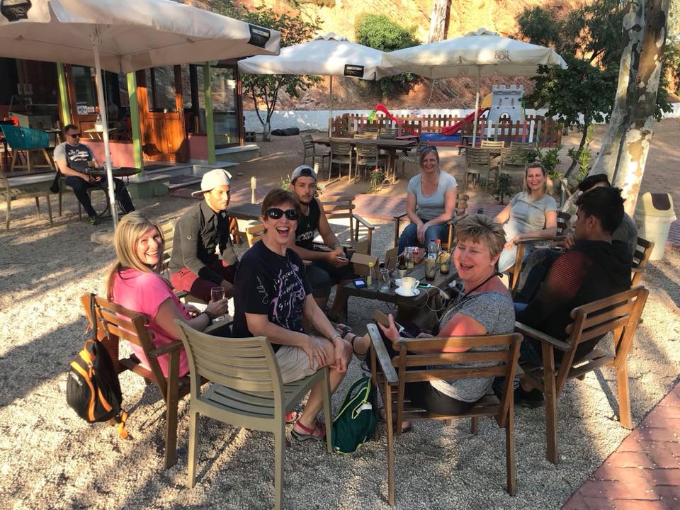On Day 2 in Chios Island, Greece, a group of people sit around a table outdoors at a cafe, smiling and talking, with drinks and bags on the table and shaded by umbrellas. | Support refugees across Europe in Greece, France, the UK and the Balkans