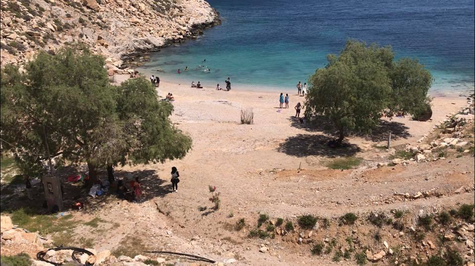 Small beach on Chios Island with people swimming and relaxing near the shore; turquoise sea, rocky terrain, and scattered trees provide shade—perfect for a peaceful Day 2 in Greece. | Support refugees across Europe in Greece, France, the UK and the Balkans