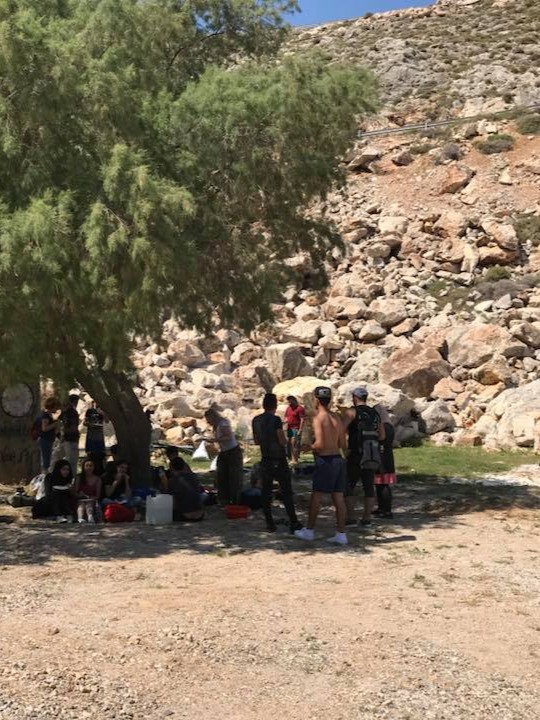 On Day 2, a group of people stands and sits under a tree for shade near rocky terrain on sunny Chios Island, Greece. | Support refugees across Europe in Greece, France, the UK and the Balkans