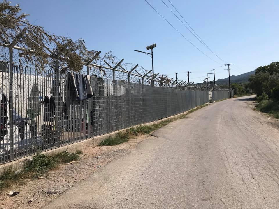 On Day 2 in Chios Island, Greece, a paved road runs alongside a tall fence topped with barbed wire. Clothes hang on the fence, and streetlights and power lines are visible under a clear sky. | Support refugees across Europe in Greece, France, the UK and the Balkans