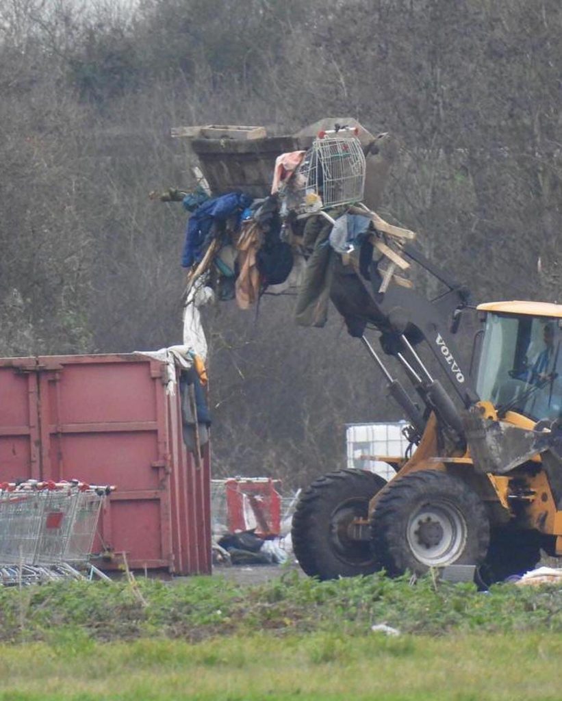 During a Police Eviction in Northern France, a Volvo backhoe loader lifts debris and a wire cage near a red dumpster, with shopping carts scattered close by and trees in the background. | Support refugees across Europe in Greece, France, the UK and the Balkans
