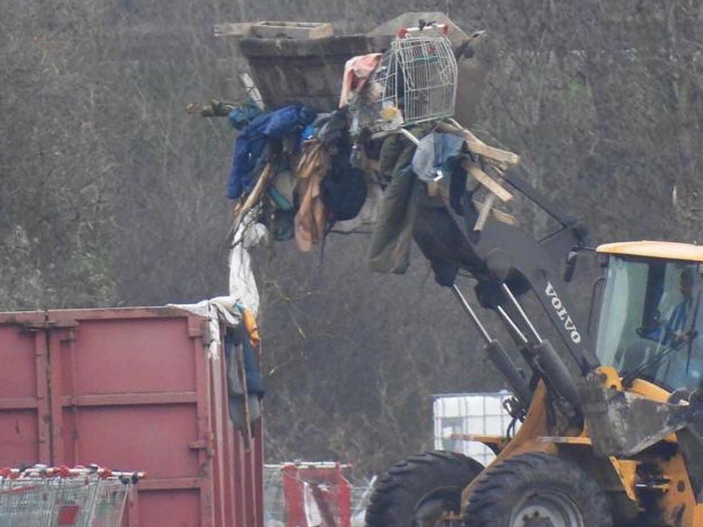 During a police eviction in Northern France, a yellow Volvo loader lifts a pile of belongings, including clothes and a metal cage, above a red container in an outdoor setting. | Support refugees across Europe in Greece, France, the UK and the Balkans