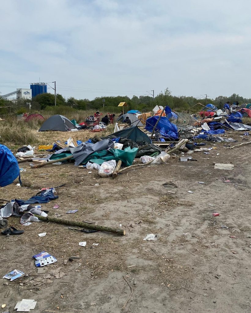 Tents and scattered debris are seen in an outdoor encampment area with litter, under a cloudy sky, near grassy terrain and industrial buildings in the background, following a recent police eviction in Northern France. | Support refugees across Europe in Greece, France, the UK and the Balkans