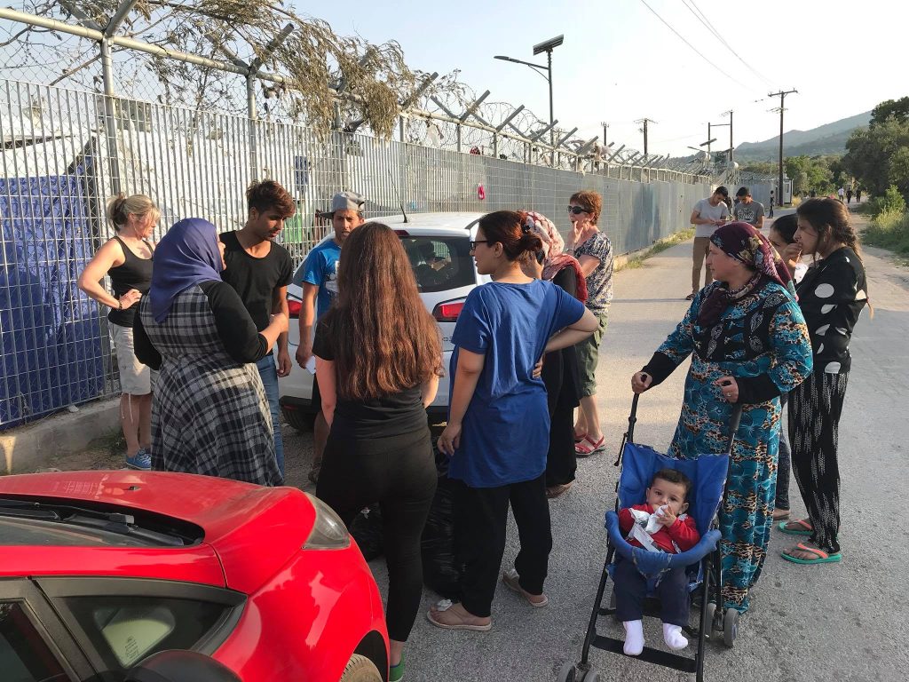 On day 3 of their travel in Chios, a group of people, including women and children, stand by parked cars near a wire fence. One woman pushes a baby in a stroller as they chat together. | Support refugees across Europe in Greece, France, the UK and the Balkans