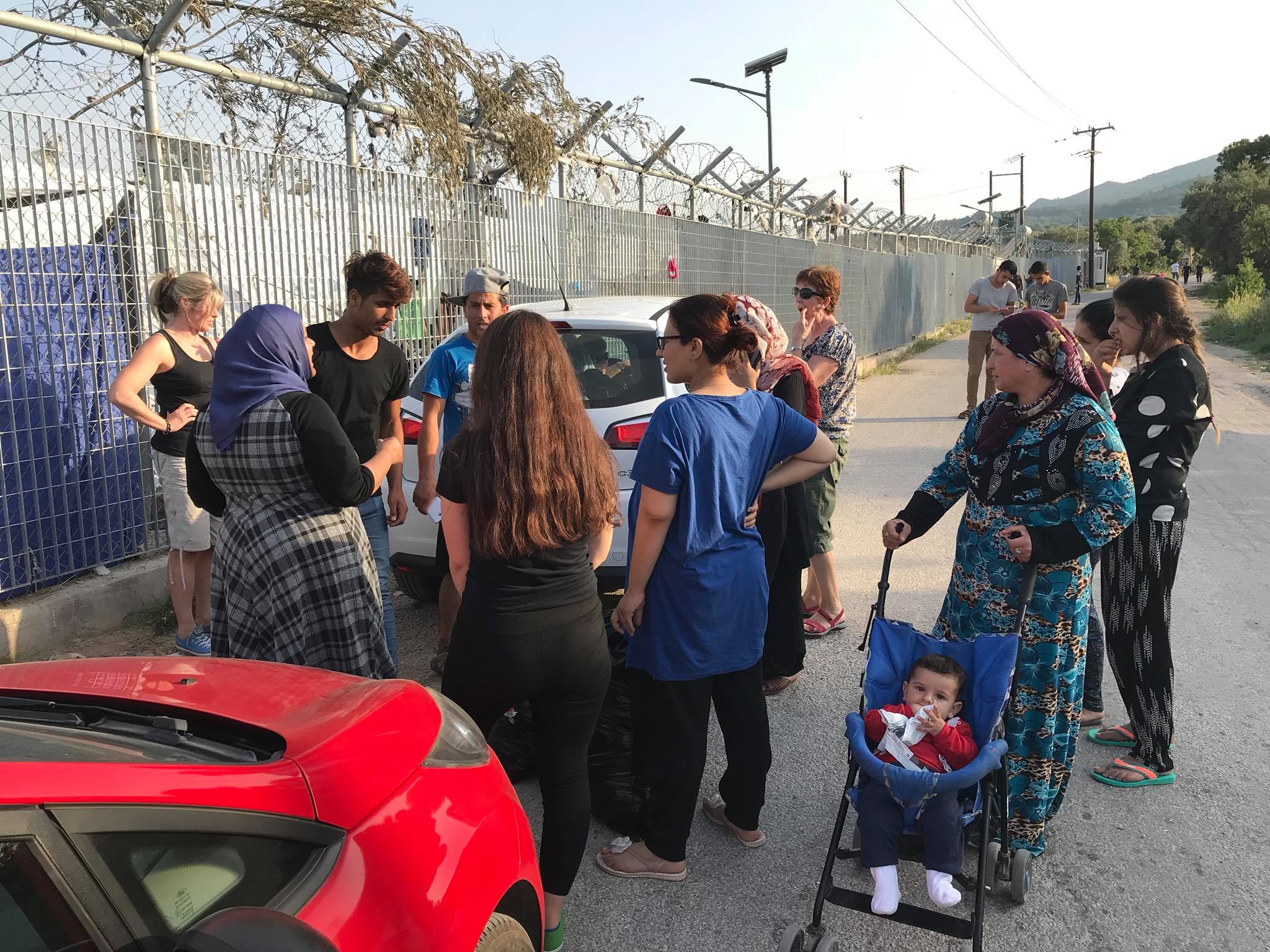 A group of people, including women and a baby in a stroller, stand and talk near parked cars beside a fenced area with barbed wire on day 3 in Chios. | Support refugees across Europe in Greece, France, the UK and the Balkans