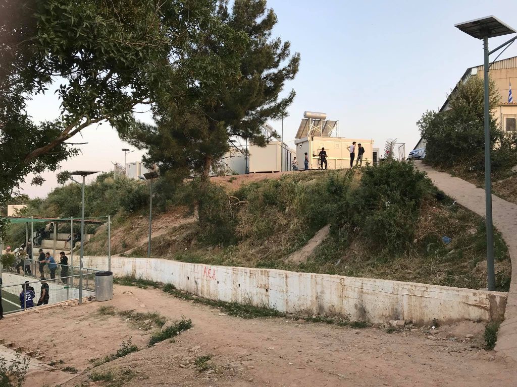 On day 3 in Chios, a sloped dirt path leads up to several portable buildings. People are walking and standing nearby, while trees and bushes are scattered around the area. | Support refugees across Europe in Greece, France, the UK and the Balkans