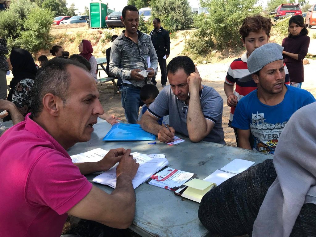 On day 3 in Chios, several adults sit at an outdoor table filling out forms and writing, while others stand nearby; trees and people are in the background. | Support refugees across Europe in Greece, France, the UK and the Balkans