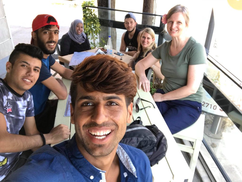 A group of seven people, men and women, are sitting around a table in a bright café in Chios on day 3 of their travel, smiling at the camera. | Support refugees across Europe in Greece, France, the UK and the Balkans