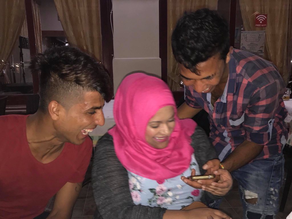 Three people sit close together in a Chios restaurant on day 3, laughing as they look at something on a smartphone. The woman in the center wears a pink hijab. | Support refugees across Europe in Greece, France, the UK and the Balkans
