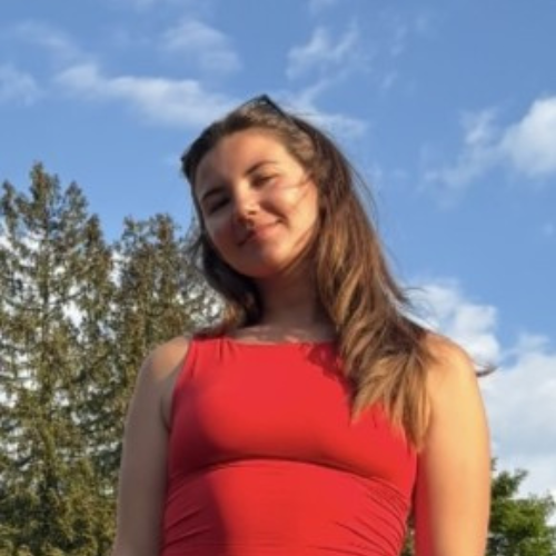 A woman in a red sleeveless top stands outdoors with trees and a blue sky in the background. | Support refugees across Europe in Greece, France, the UK and the Balkans