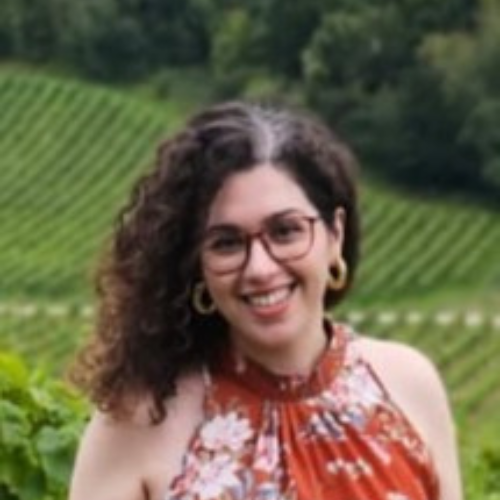 A woman with curly hair and glasses smiles at the camera, standing outdoors in front of green, terraced fields. | Support refugees across Europe in Greece, France, the UK and the Balkans