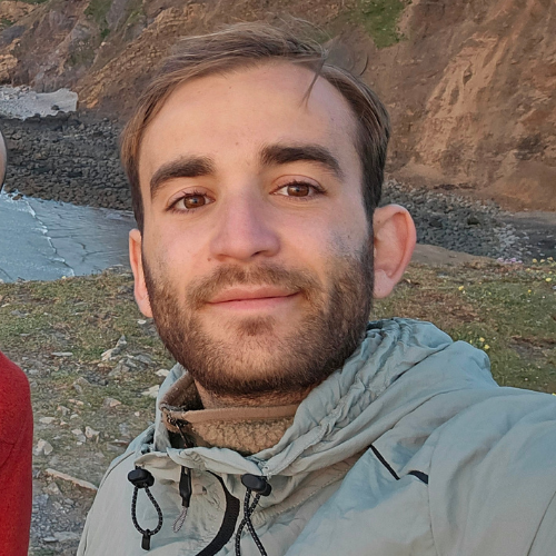 A man with short brown hair and a beard wears a light gray jacket and stands outdoors with rocky cliffs and the ocean in the background. | Support refugees across Europe in Greece, France, the UK and the Balkans