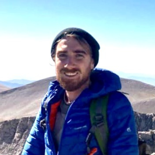 Man wearing a blue jacket and black beanie stands outdoors with mountains in the background on a clear day. | Support refugees across Europe in Greece, France, the UK and the Balkans