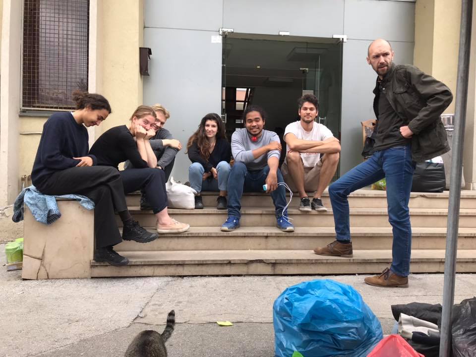 Seven people sit and stand on steps outside a building in Sarajevo, BiH, with bags and a cat visible in the foreground. They appear relaxed and are facing the camera on Day 6 of their journey. | Support refugees across Europe in Greece, France, the UK and the Balkans