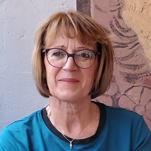 A woman with short, light brown hair and glasses, wearing a blue top, sits in front of a textured wall with abstract patterns. | Support refugees across Europe in Greece, France, the UK and the Balkans