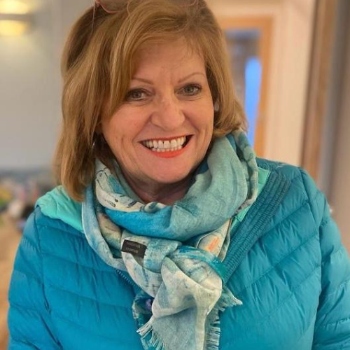 A woman with shoulder-length light brown hair smiles, wearing a blue jacket and a patterned blue scarf indoors. | Support refugees across Europe in Greece, France, the UK and the Balkans
