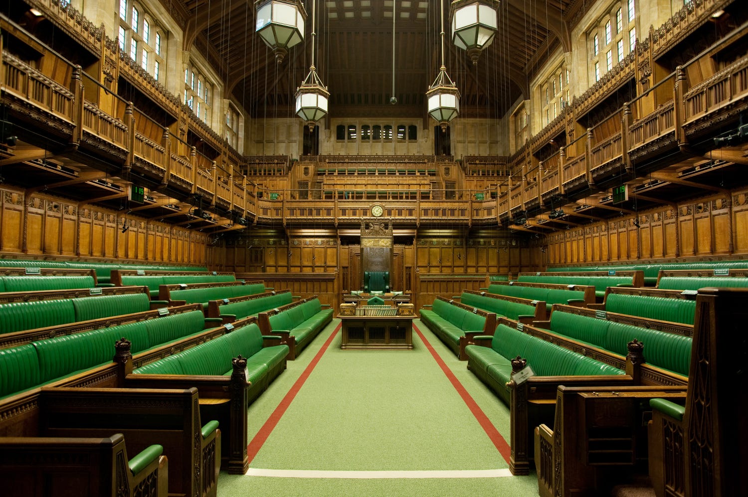 The interior of the House of Commons in the UK Parliament features green benches, wooden paneling, a central table, and the speaker's chair at the far end—a place where you can write to your MP to stand with refugees and support people seeking safety. | Support refugees across Europe in Greece, France, the UK and the Balkans