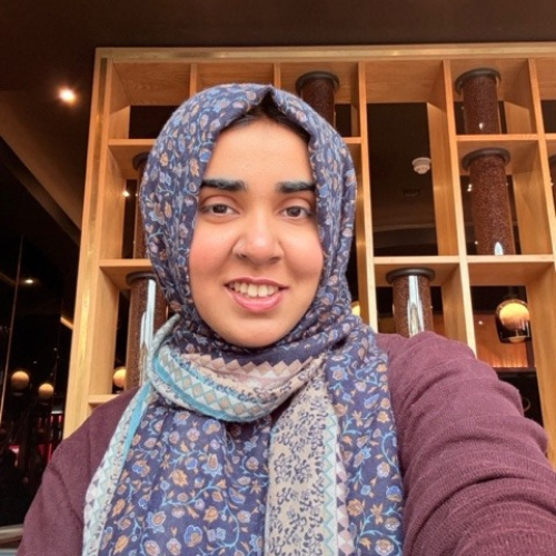 A person wearing a patterned hijab and maroon sweater smiles at the camera, sitting indoors with wooden shelves and cylindrical decor in the background. | Support refugees across Europe in Greece, France, the UK and the Balkans