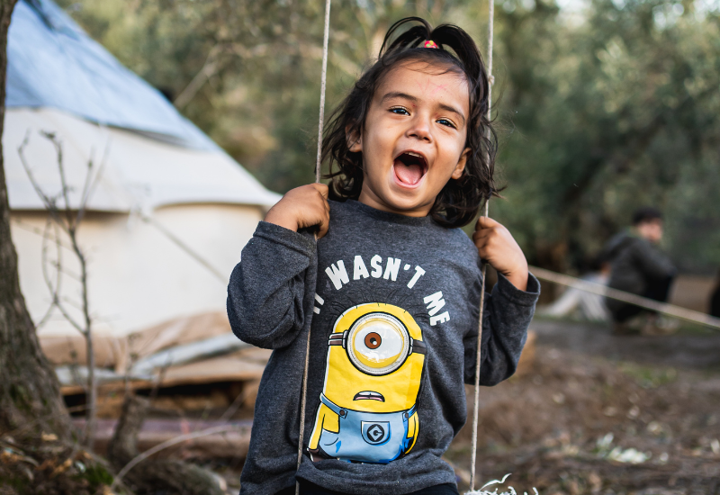 Young child with dark hair and a ponytail smiles widely while sitting on a swing, wearing a shirt with a cartoon minion and the text "IT WASN'T ME." Outdoor setting with trees and a tent. | Support refugees across Europe in Greece, France, the UK and the Balkans