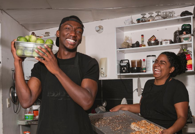 Two people in a kitchen wear black aprons and smile; one holds a container of green apples, the other holds a tray with baked goods. Shelves with kitchen items are visible in the background. | Support refugees across Europe in Greece, France, the UK and the Balkans
