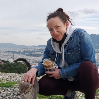 A woman in a denim jacket crouches and pets a cat outdoors, holding a drink, with a cityscape and mountains in the background. | Support refugees across Europe in Greece, France, the UK and the Balkans