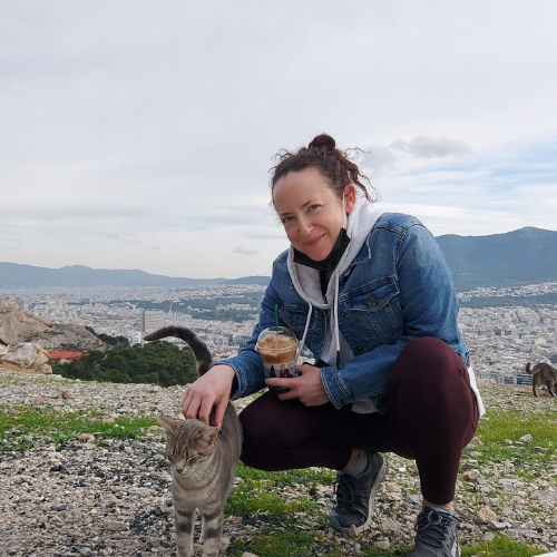 A woman in a denim jacket crouches and pets a cat outdoors, holding a drink, with a cityscape and mountains in the background. | Support refugees across Europe in Greece, France, the UK and the Balkans