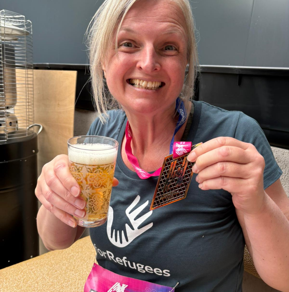A smiling woman holds a glass of beer in one hand and a geometric medal in the other. She wears a shirt with a "ForRefugees" logo and a colorful lanyard around her neck. | Support refugees across Europe in Greece, France, the UK and the Balkans