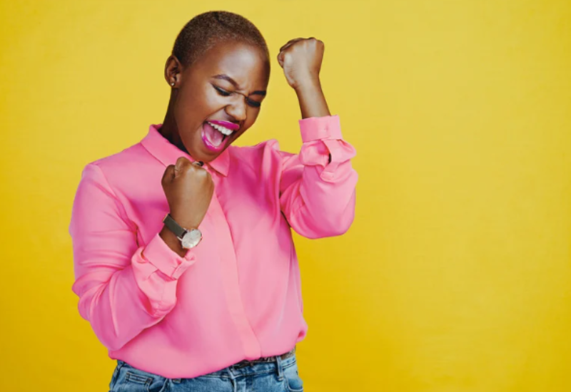 A person wearing a bright pink shirt and blue jeans smiles with eyes closed and raises both fists in a celebratory gesture against a yellow background. | Support refugees across Europe in Greece, France, the UK and the Balkans