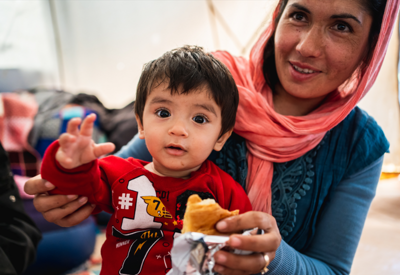 A woman in a pink headscarf holds a young child wearing a red shirt, who is reaching out and holding a piece of bread. | Support refugees across Europe in Greece, France, the UK and the Balkans