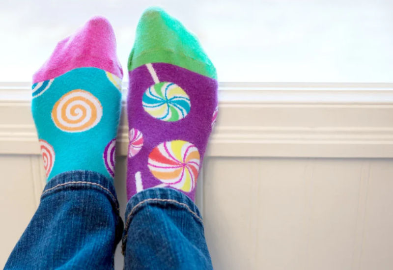 A person wearing mismatched, colorful candy-patterned socks and blue jeans sits with feet propped up on a white windowsill. | Support refugees across Europe in Greece, France, the UK and the Balkans
