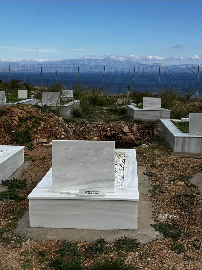 A white marble grave with a headstone and small plaque rests in a cemetery on a hill, overlooking the sea and mountains under a partly cloudy sky—a quiet tribute to stories of strength, hope and love, perfect for Mother’s Day remembrance. | Support refugees across Europe in Greece, France, the UK and the Balkans