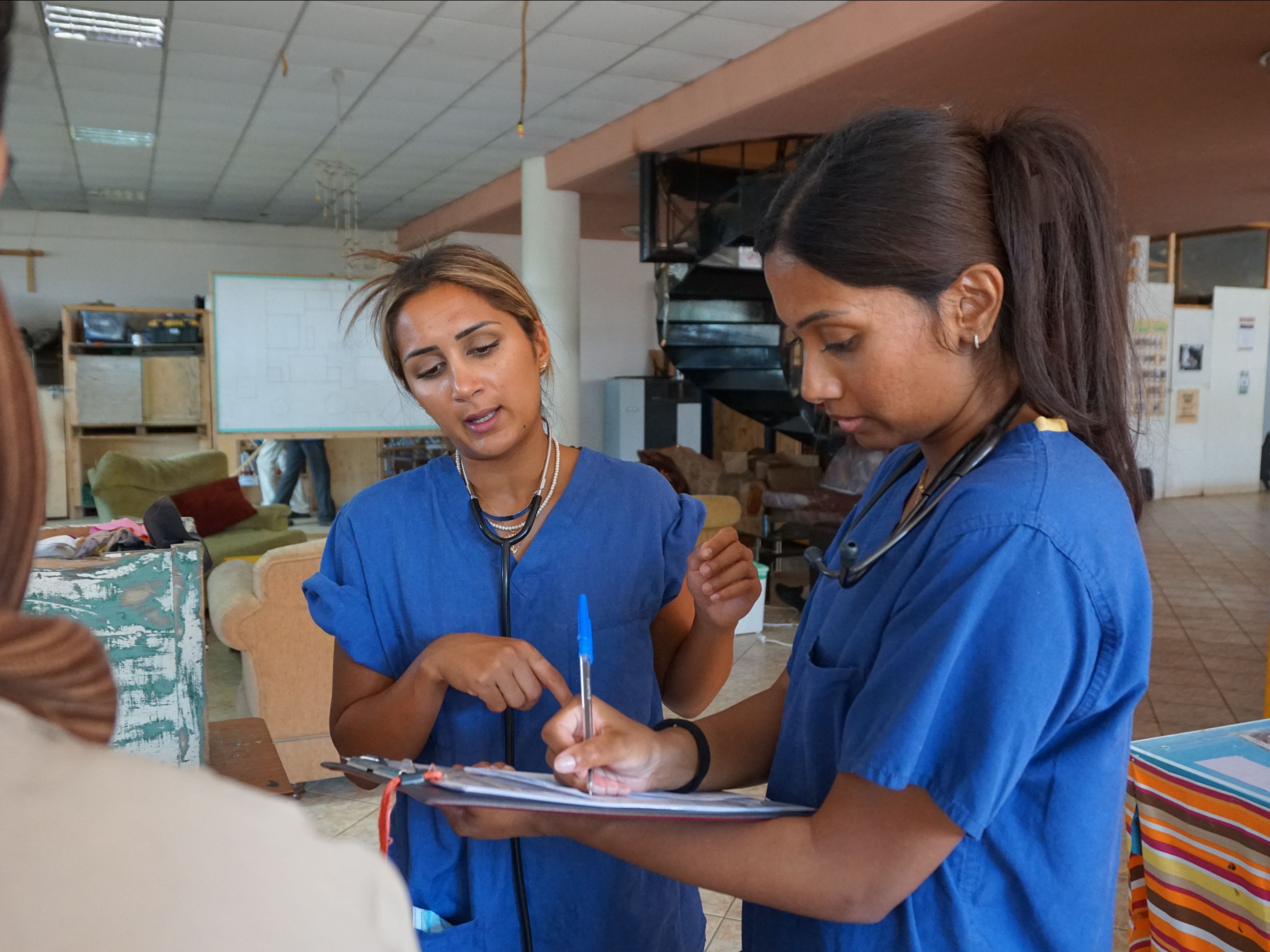 Two women in blue scrubs stand indoors; one gestures while speaking, and the other writes notes on a clipboard, reflecting their dedication to community healthcare. | Support refugees across Europe in Greece, France, the UK and the Balkans