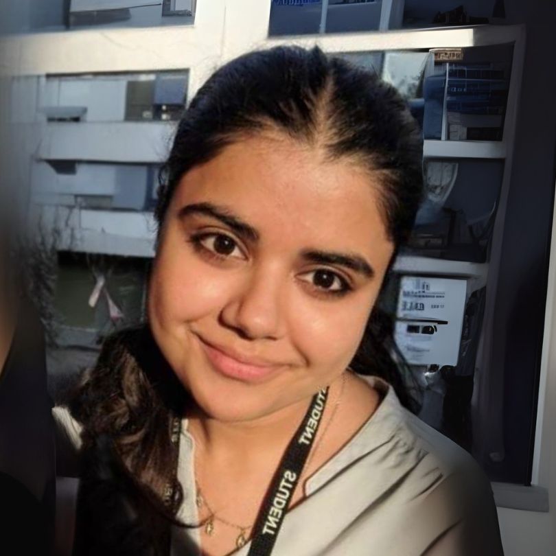 A woman with dark hair smiles at the camera, wearing a light-colored top and a lanyard, with buildings and windows visible in the background. | Support refugees across Europe in Greece, France, the UK and the Balkans