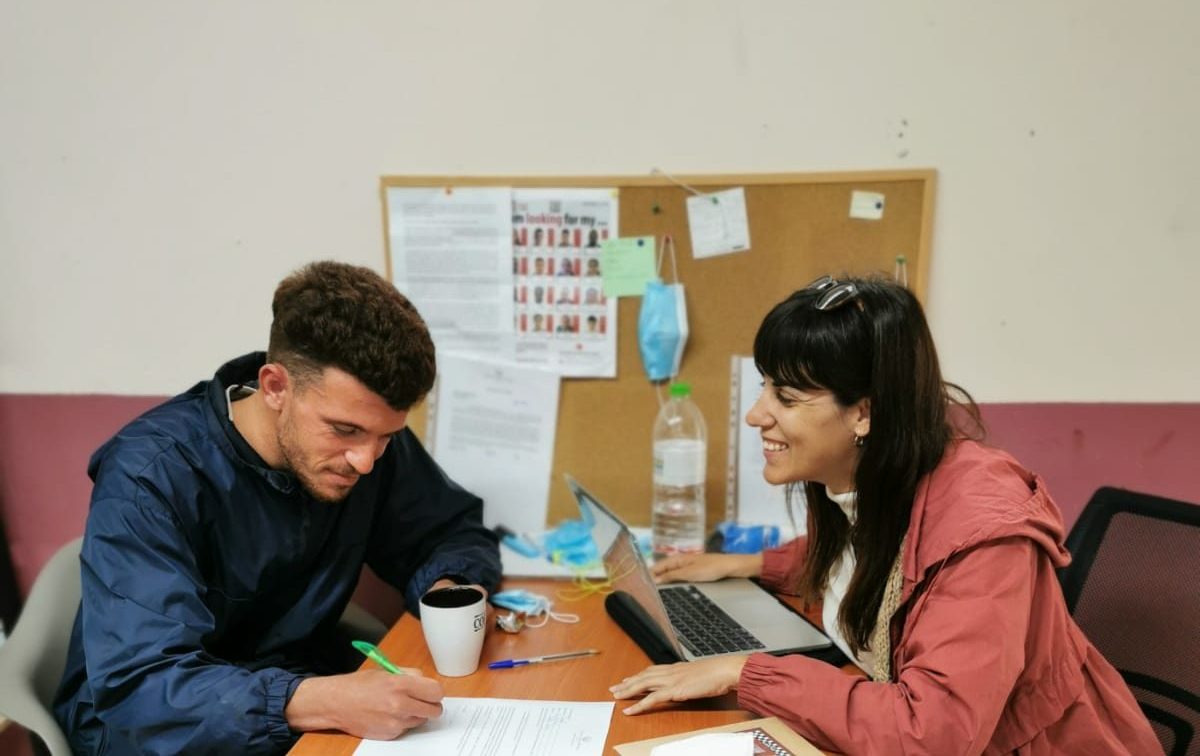 Two people sit at a desk, reviewing and signing a document about stranded refugees. A laptop, books, and coffee cup are on the table; a bulletin board is in the background. | Support refugees across Europe in Greece, France, the UK and the Balkans