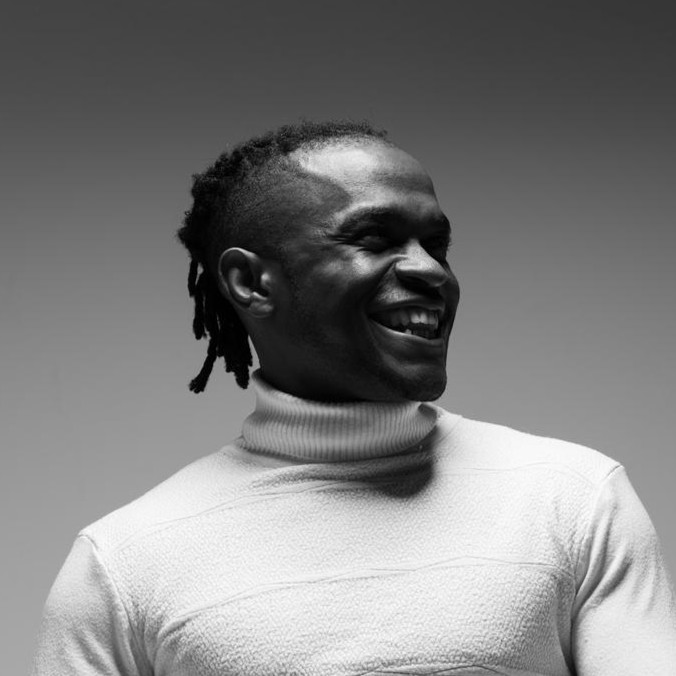 A man wearing a light-colored turtleneck sweater smiles and looks to the side against a plain background. The photo is in black and white. | Support refugees across Europe in Greece, France, the UK and the Balkans