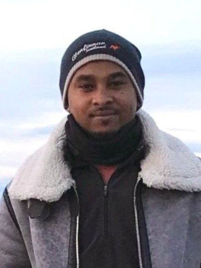 A man wearing a black beanie and a gray jacket with a shearling collar stands outdoors in front of a cloudy sky, embodying stories of strength and hope and love. | Support refugees across Europe in Greece, France, the UK and the Balkans