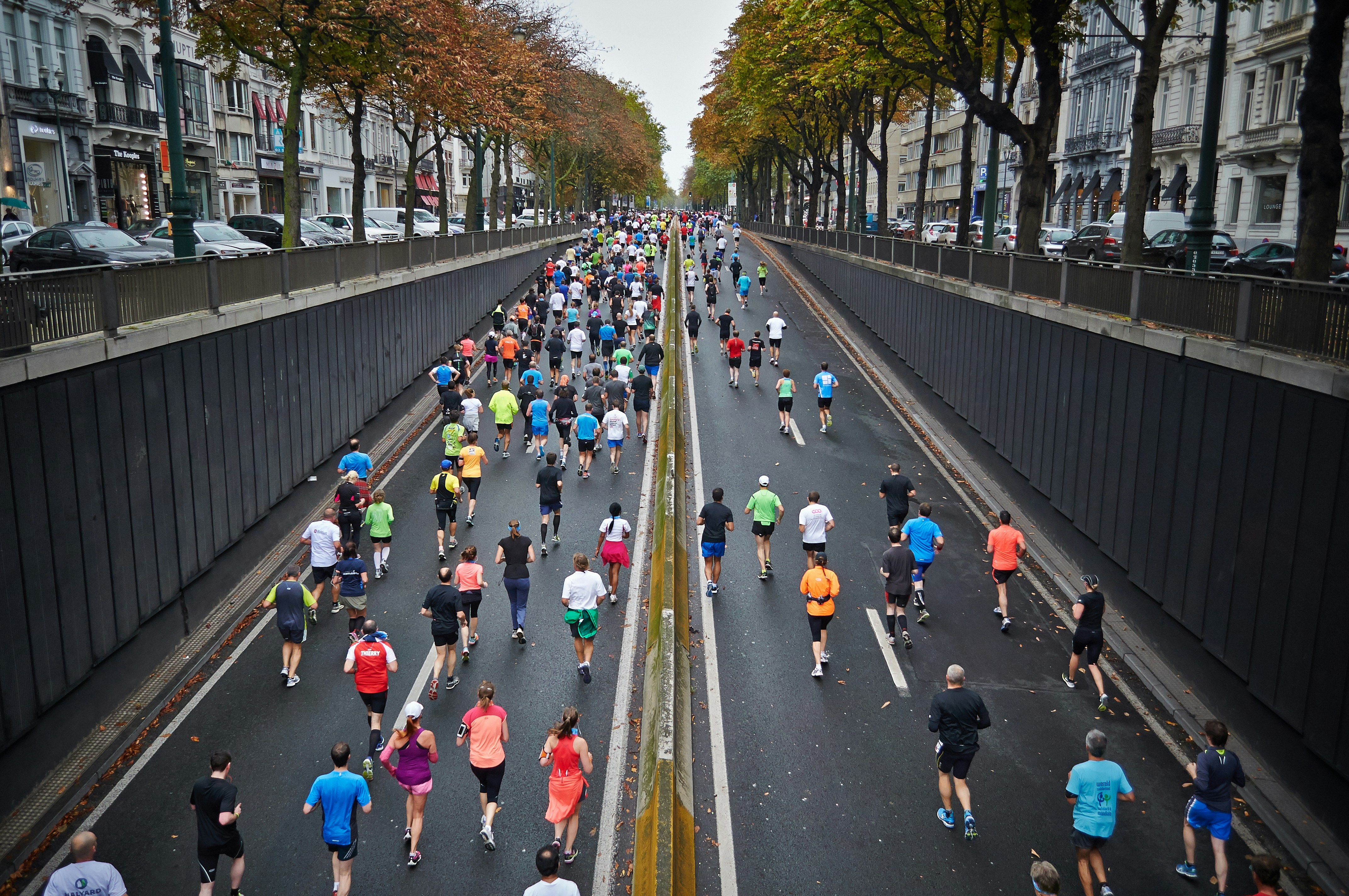 A large group of runners participates in a road race on a city street lined with trees and buildings, viewed from above. | Support refugees across Europe in Greece, France, the UK and the Balkans