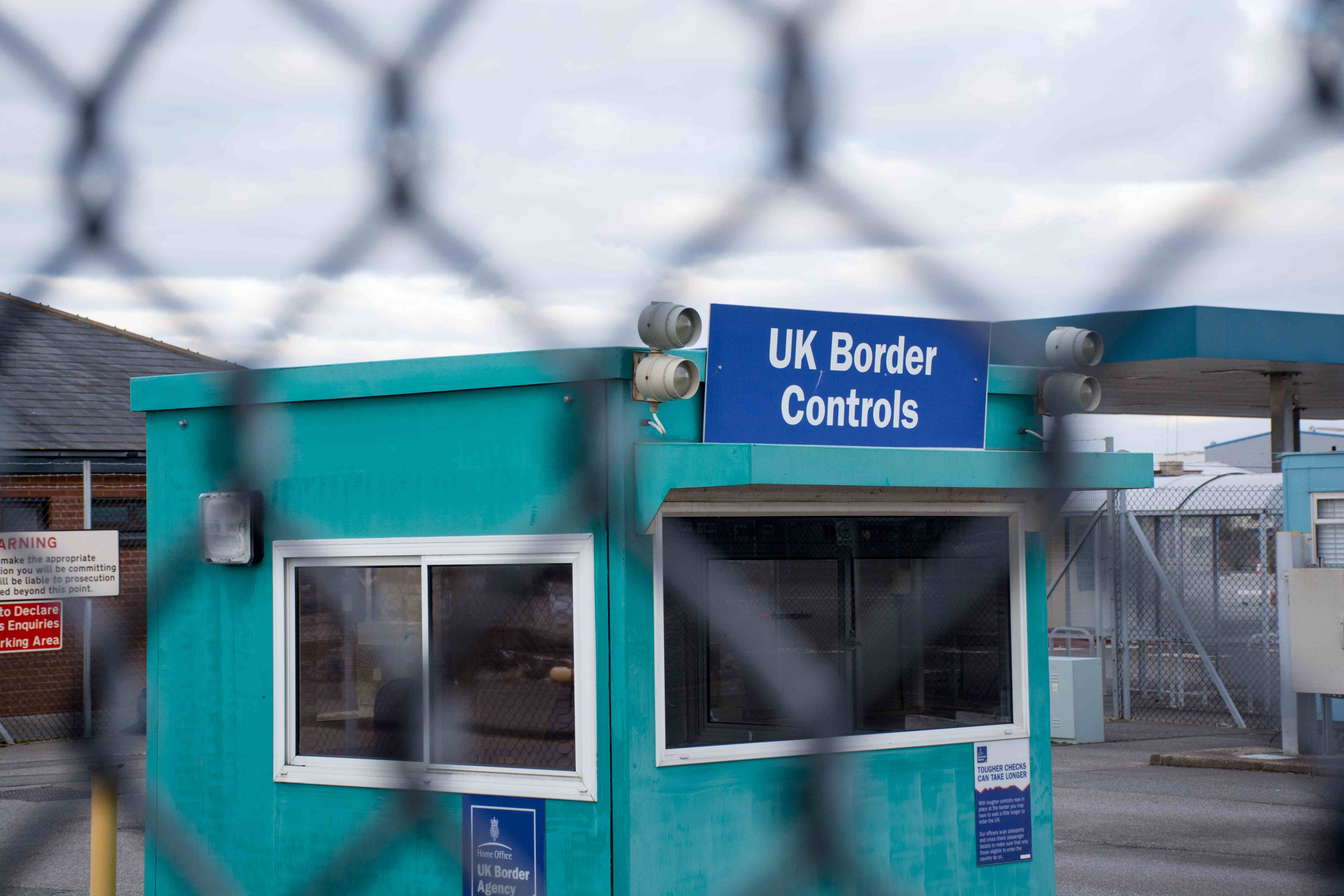 A small blue border control booth with a "UK Border Controls" sign, security cameras, and fencing, viewed through a chain-link fence. | Support refugees across Europe in Greece, France, the UK and the Balkans